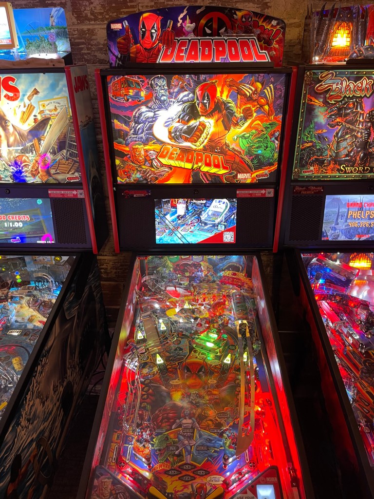 A close-up view of a 'Deadpool' pinball machine featuring vibrant artwork and illuminated playfield in Cherry Lanes Arcade Bar.
