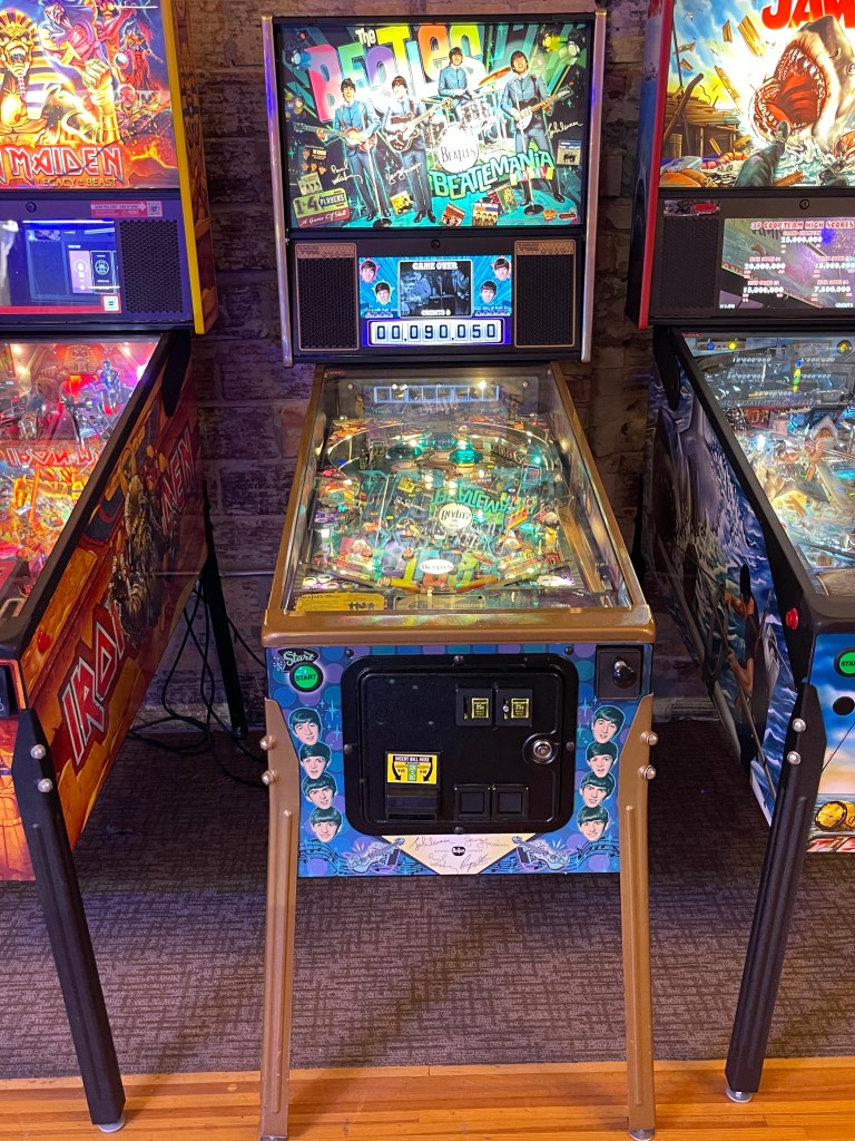 A close-up view of a Beatles-themed pinball machine, showing the colorful playfield and controls, located in an arcade setting with other pinball machines in the background.